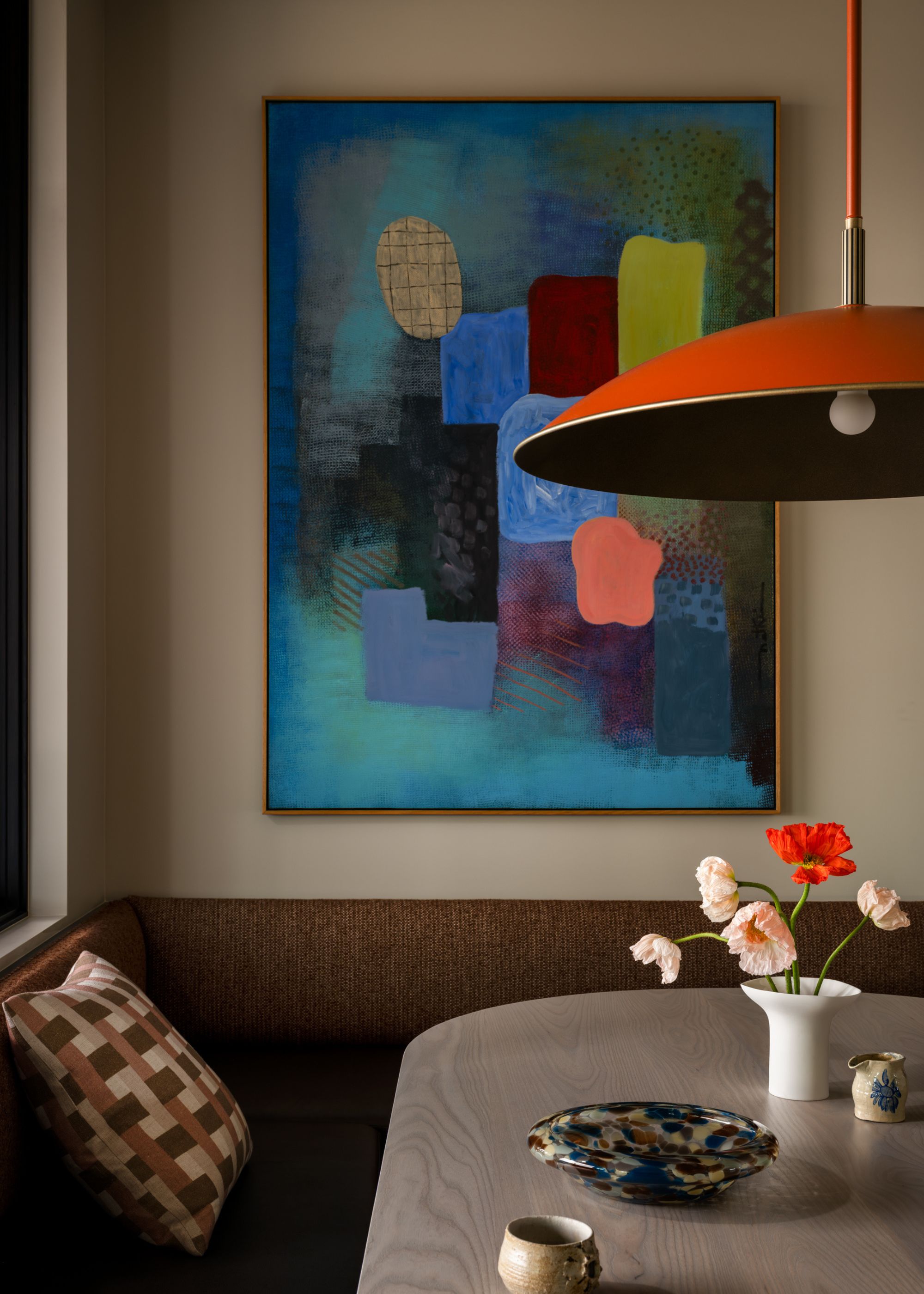 A banquette seating area with colorful artwork above it and a red pendant light above the table with a decorative tray and a small vase with pink and red floral stems
