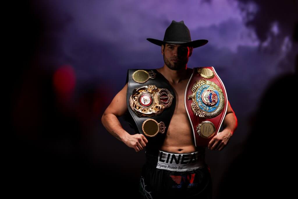 Gilberto "Zurdo" Ramirez posing shirtless with two belts and a cowboy hat.