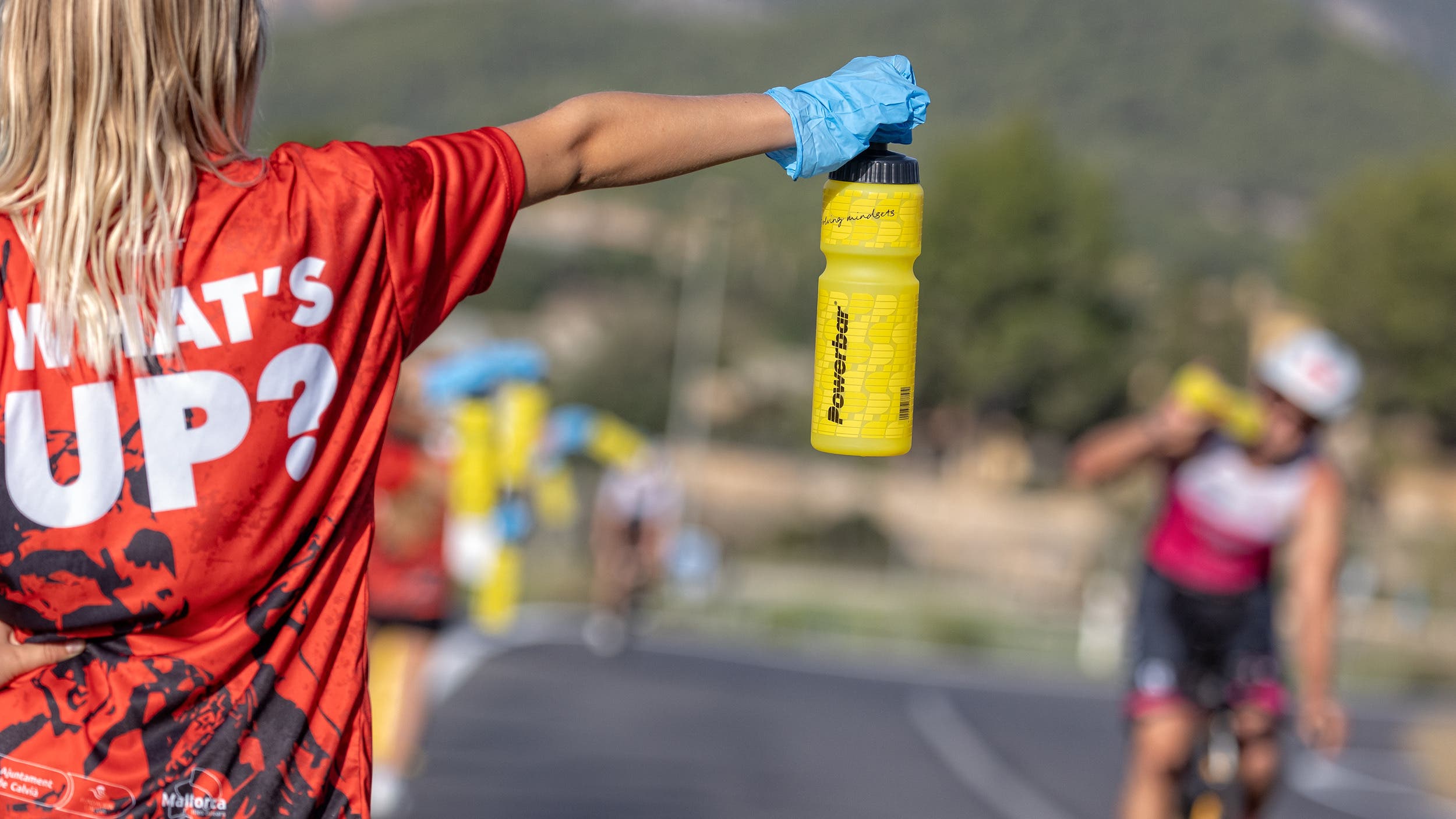 An aid station volunteer holds out a bottle of sports drink to a triathlete on a bike.