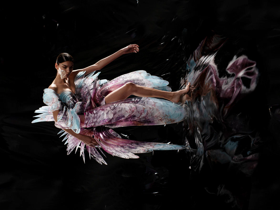 Woman in an artistic, flowing dress with feather-like textures, set against a dark, abstract background.
