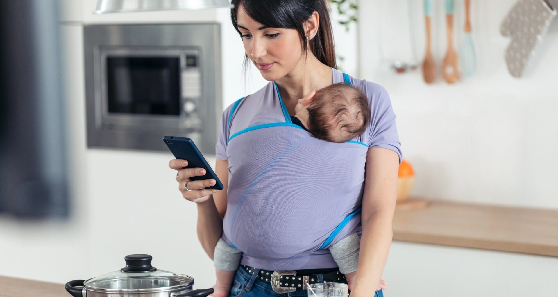Shot of pretty young mother with little baby in sling using her mobile phone in the kitchen at home.