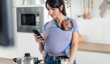 Shot of pretty young mother with little baby in sling using her mobile phone in the kitchen at home.
