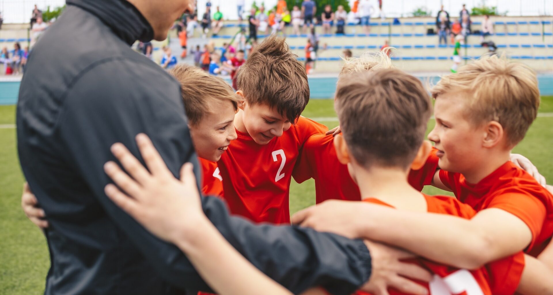 Happy boys in sports team huddling with young coach. Kids standing in a circle on grass sports pitch. Children sports team