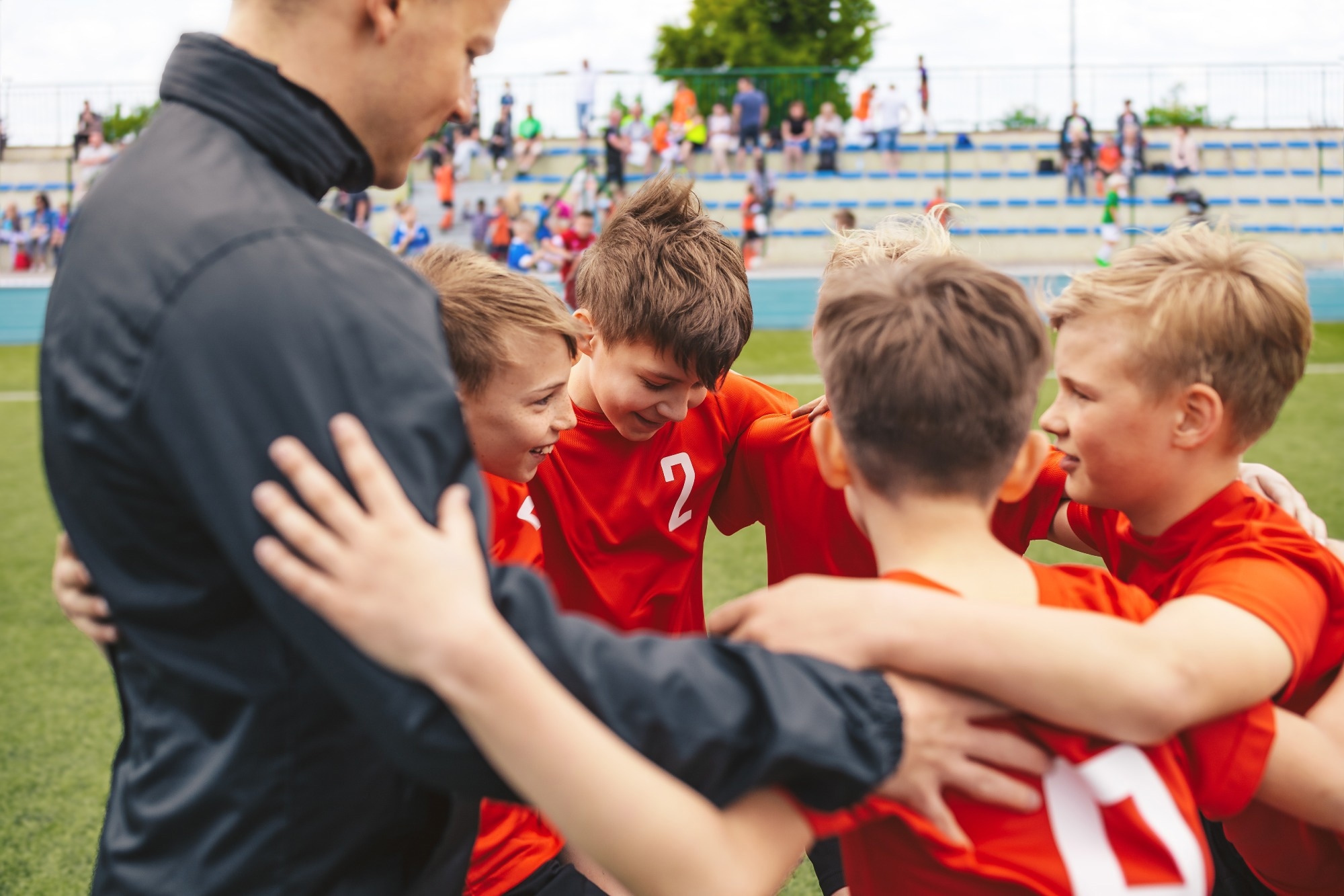 Happy boys in sports team huddling with young coach. Kids standing in a circle on grass sports pitch. Children sports team