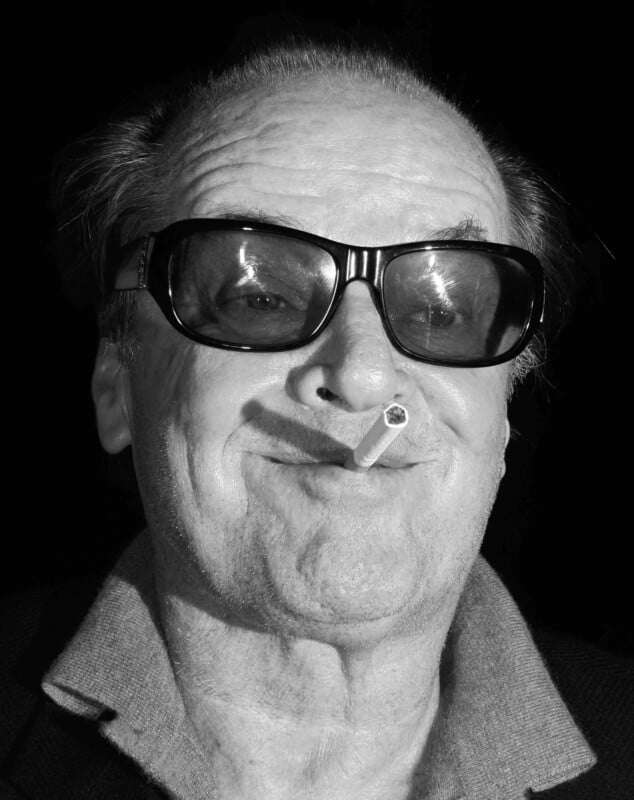 Older man with receding hairline wears large dark sunglasses and a collared shirt, smirking with a cigarette held between his lips. The background is dark and the image is black and white.