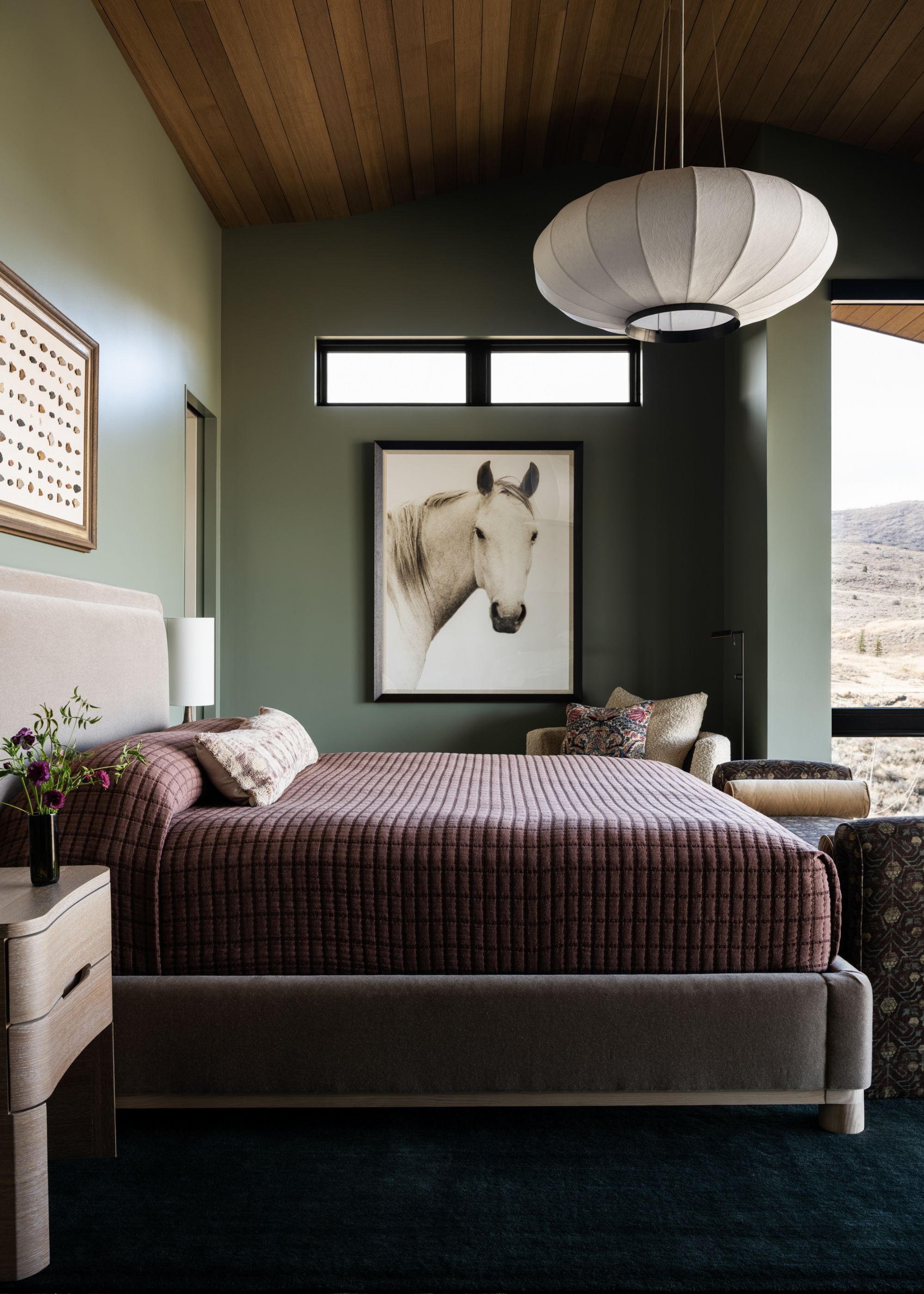 A contemporary bedroom with green walls and wood paneled ceiling with a framed picture of a horse below two rectangular windows that pairs with the white pendant light above the bed
