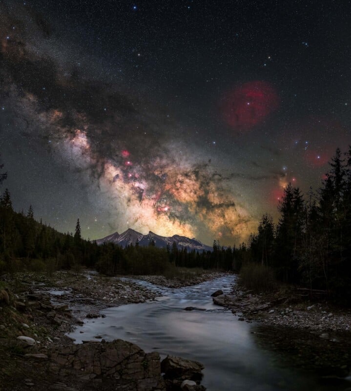 A star-filled night sky with the bright, colorful Milky Way galaxy stretches above a forested river, mountains silhouetted on the horizon, and rocky riverbanks in the foreground.