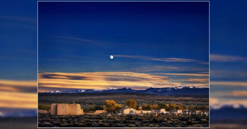 Adobe church and graveyard under a twilight sky with a full moon and snow-capped mountains in the distance.