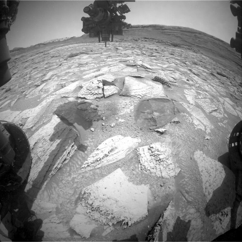 Black and white image taken by a Mars rover showing rocky terrain with flat, cracked slabs in the foreground. Parts of the rover are visible on the sides and top, with a distant ridge under a hazy sky in the background.