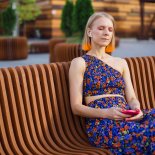 Relaxed young woman in bright summer clothes is sitting on a bench on a city street with her eyes closed enjoying music through headphones from a smartphone.