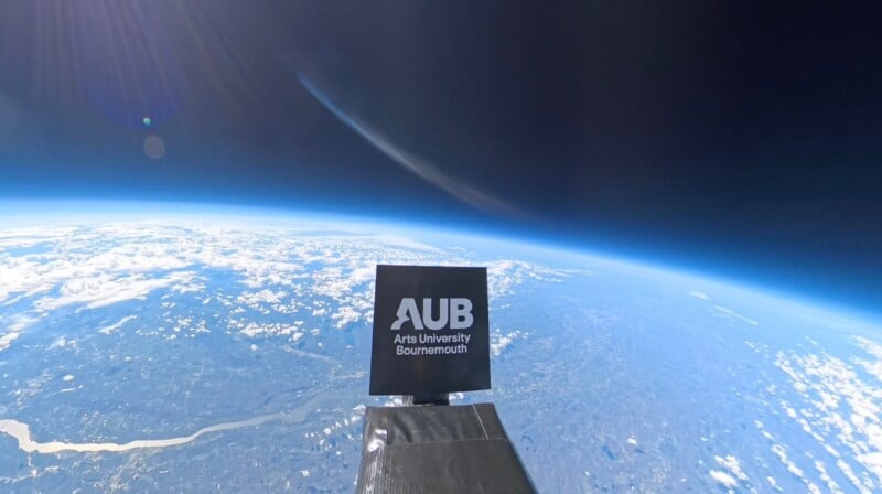 A sign reading "AUB Arts University Bournemouth" is attached to a device floating high above the Earth, with the planet’s curved horizon, blue atmosphere, scattered clouds, and land visible below.