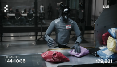 A humanoid robot with a gray body and black, reflective head stands next to a package sorting station while moving boxes and padded envelopes onto a conveyor belt.