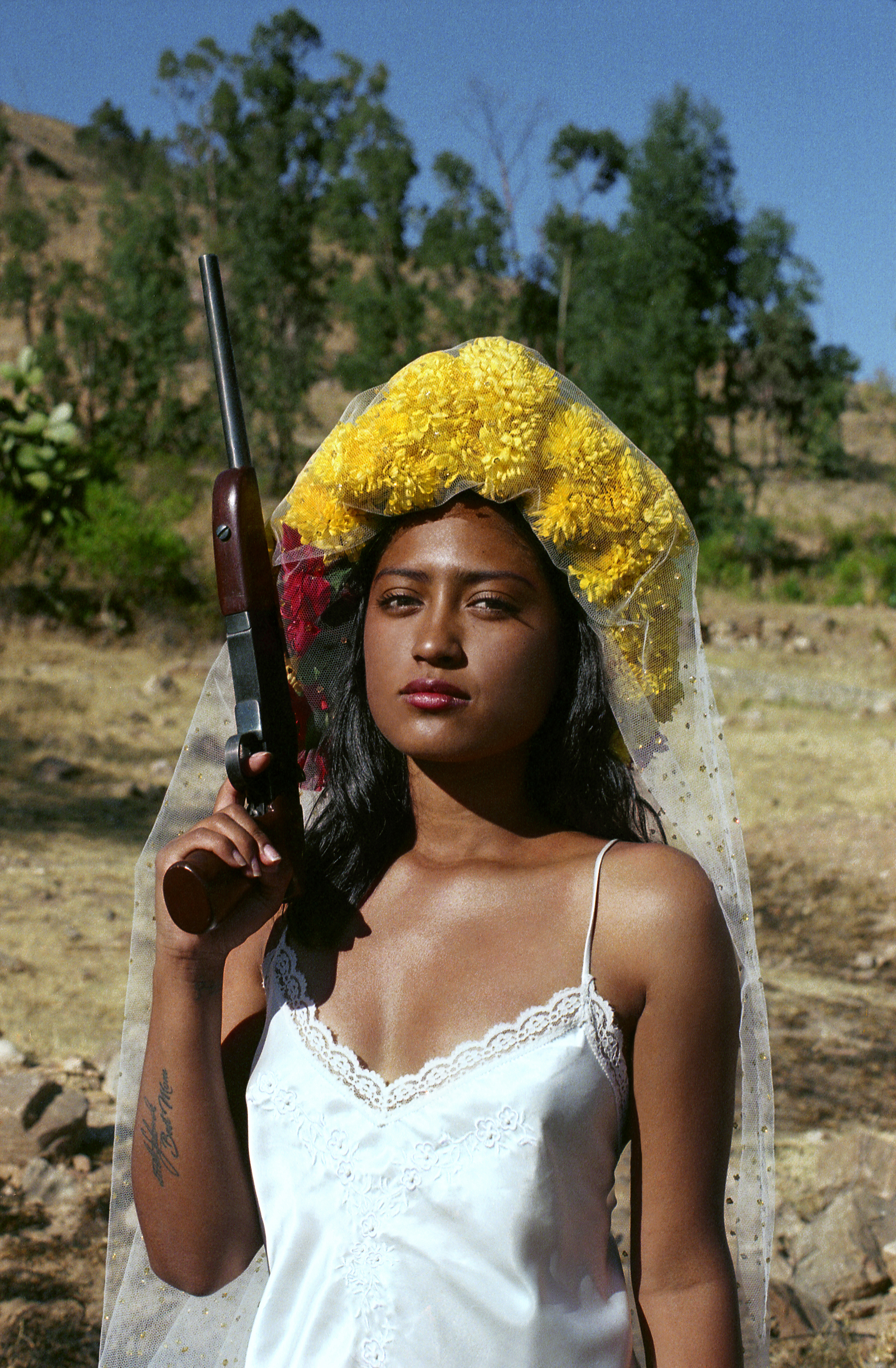 A woman in a white slip dress and a veil covered in yellow flowers holds a shotgun upright at her shoulder, standing in a dry, rocky landscape under a clear sky.