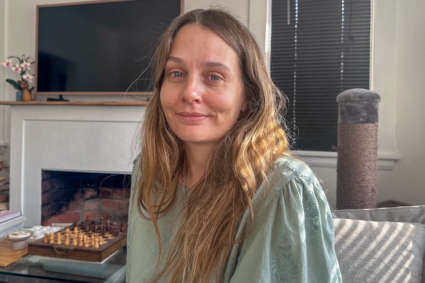 A woman with long, fair hair and a pale green top sits in a home.