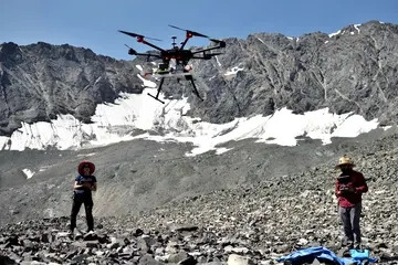 Two people standing on a rocky surface with mountains in the background. The person on the right is holding a controller, controlling the drone in the middle of them above the ground.