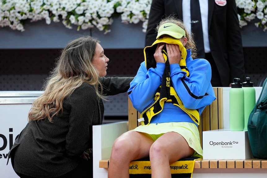 Female tennis player cries into a towel while seated courtside as a woman in black clothing consoles her. 