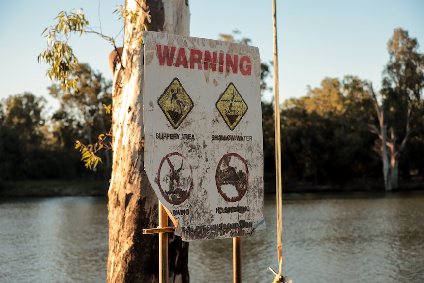 A faded sign sits in front of a gum tree with water behind. It reads WARNING SLIPPERY AREA SHALLOW WATER NO DIVING NO SWIMMING