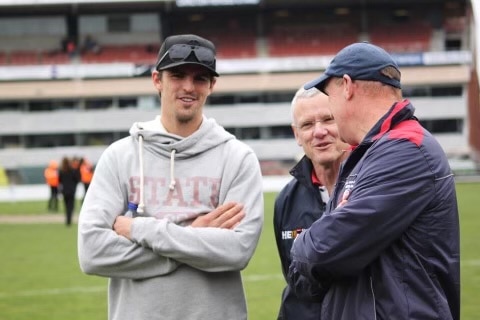 A young man in a cap and hoodie folds his arms while standing with two older men a stadium football ground.