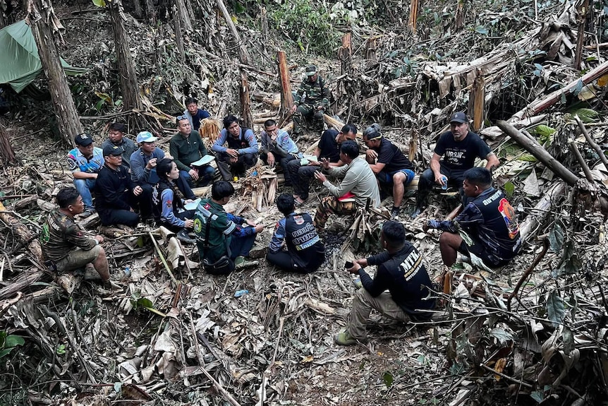 About a dozen people sit in a circle in the jungle listening to a person talking.