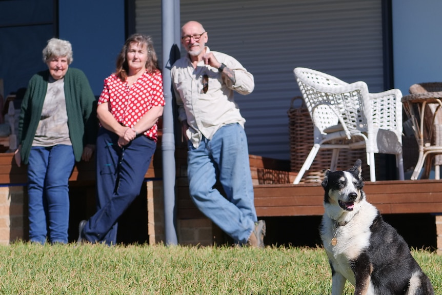 Three people in background leaning against deck, dog in foreground.