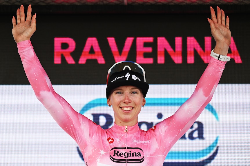 Lorena Wiebes smiles and holds up her hands while wearing a pink leader's jersey.