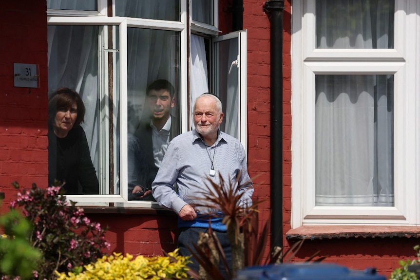 A woman and a man peer out a window, looking concerned. Another man stands near the window, looking on.