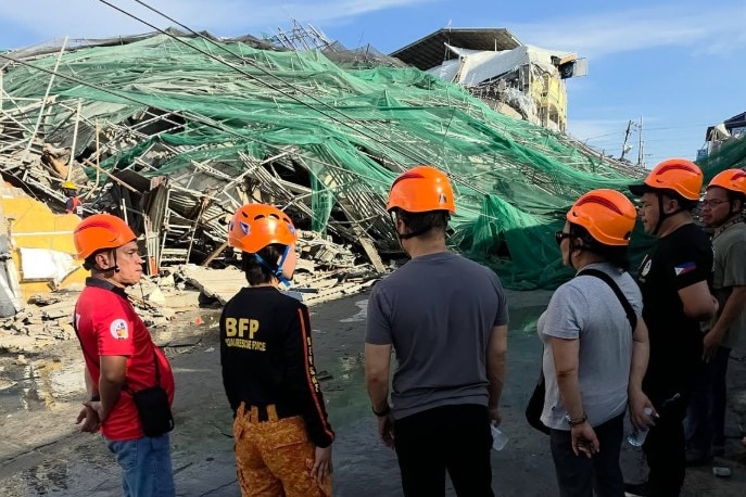 A group of people wearing protective hats stand in front of the collapsed building.