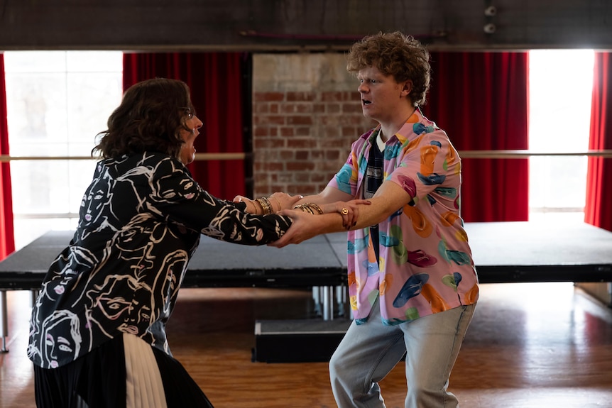 Anne and Will hold onto each other's arms while standing in a theatre rehearsal space and yelling.