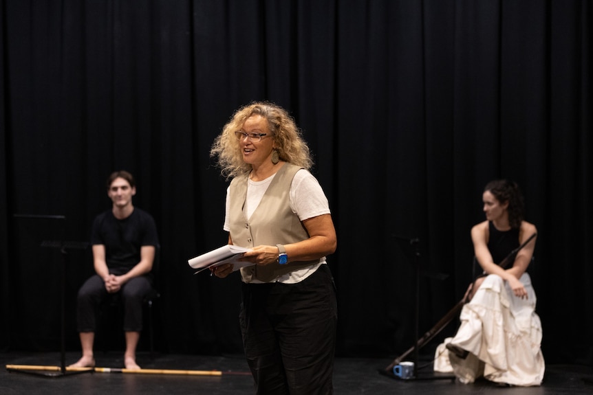 Leah Purcell holds a book and talks to cast members at a rehearsal. Cast members are out of focus in the background.