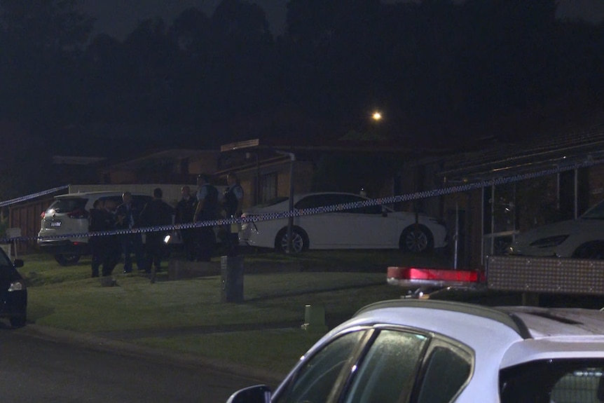 Police and investigators in a group on the front lawn of a house surrounded by police tape.