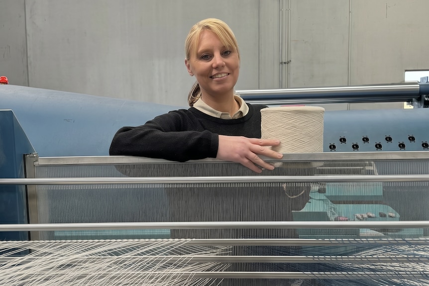 A young blonde woman stands in front of a industrial size loom holding a cream cololured spool of yarn, she's smiling