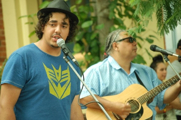 Goori, wears a hat, sings into a mic, his dad Buddy beside him playing guitar, greenery, another woman behind them.