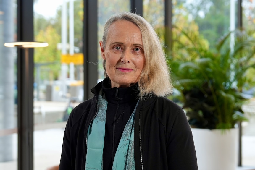 A woman with a blonde bob stands indoors looking serious.