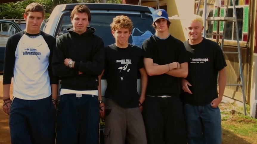 Five young men, possibly teenagers, stand leaning against a car. One has arms crossed