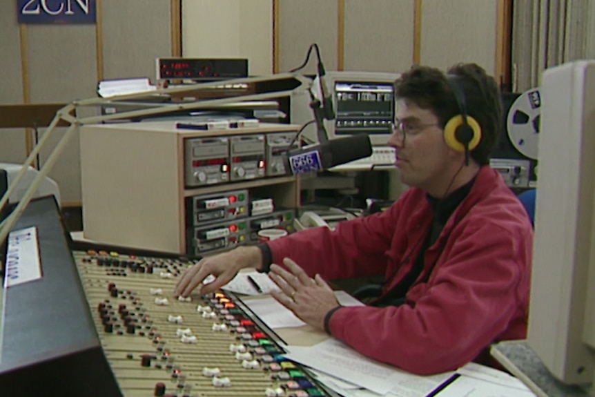 James Valentine wears a long-sleeve red shirt, glasses and a headset as he moves buttons on a radio panel board