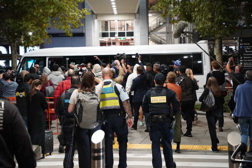 A large group of police and people, including people holding mobile phones and tv cameras, in a tight scrum near a white bus.