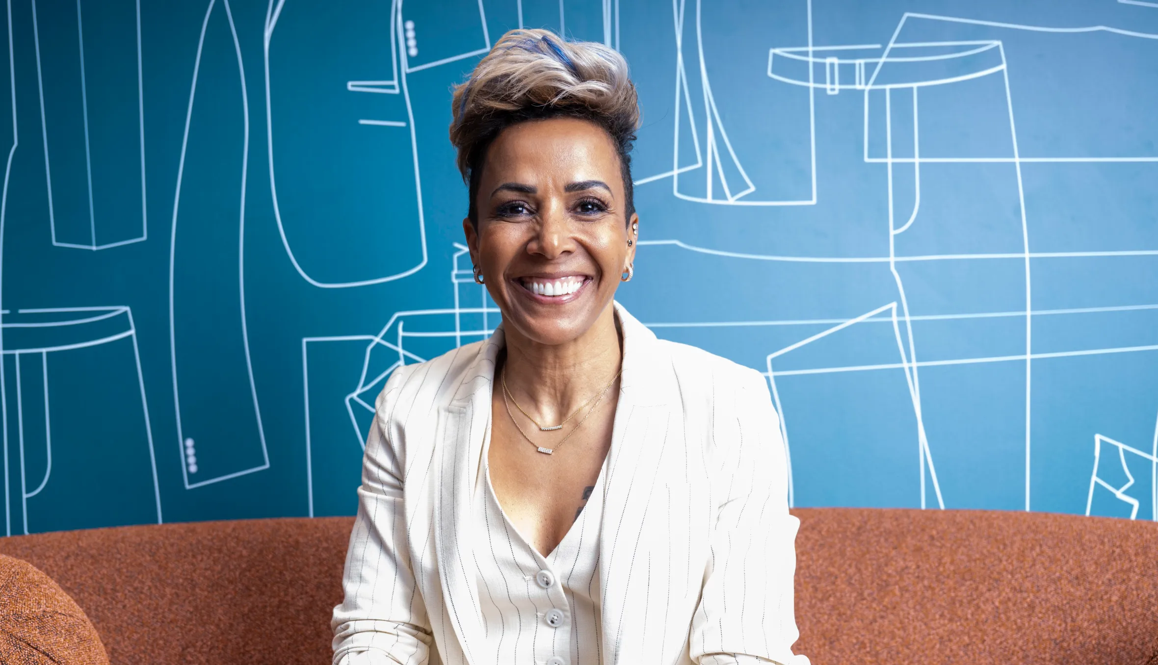 Dame Kelly Holmes, in a cream pinstriped suit, smiling while seated on an orange couch.