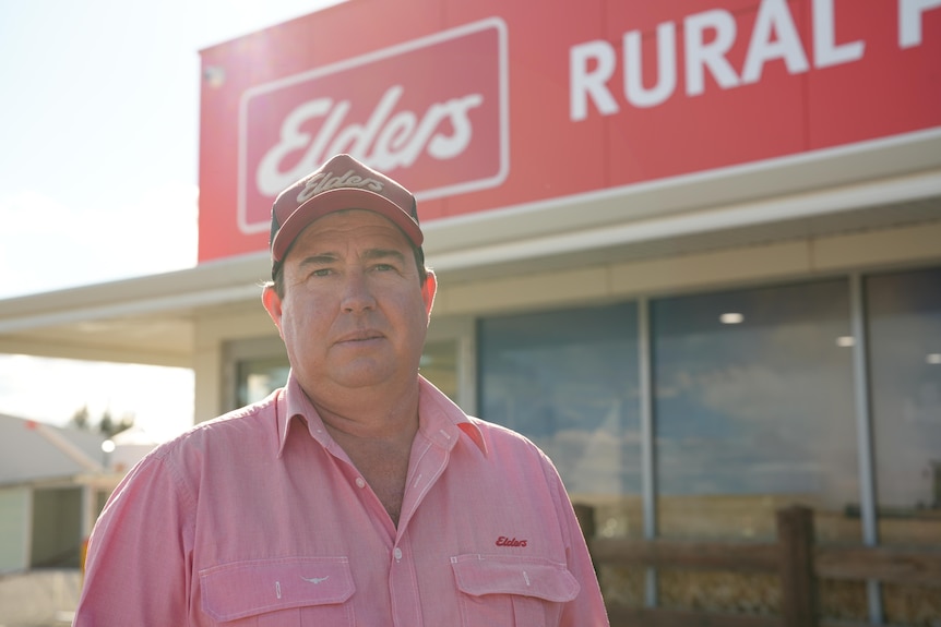 A man with a pink shirt and red hat looking at the camera with a red building behind him.