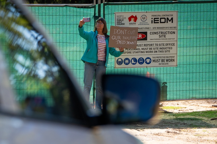 A woman holds a sign saying "Don't cut our beautiful trees down!" as a ute approaches a gate
