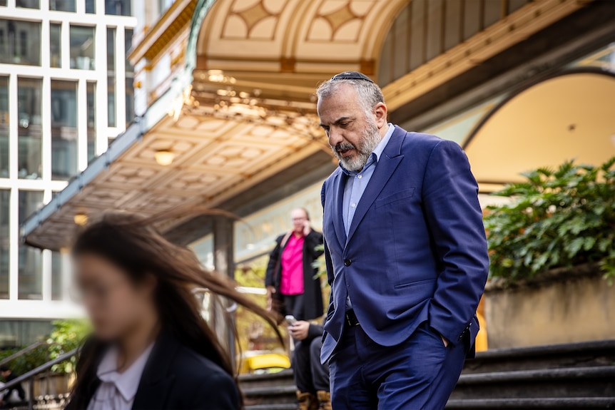 A bearded man with grey hair wears a yarmulke and a dark suit as he ascends a flight stairs outside a court building.