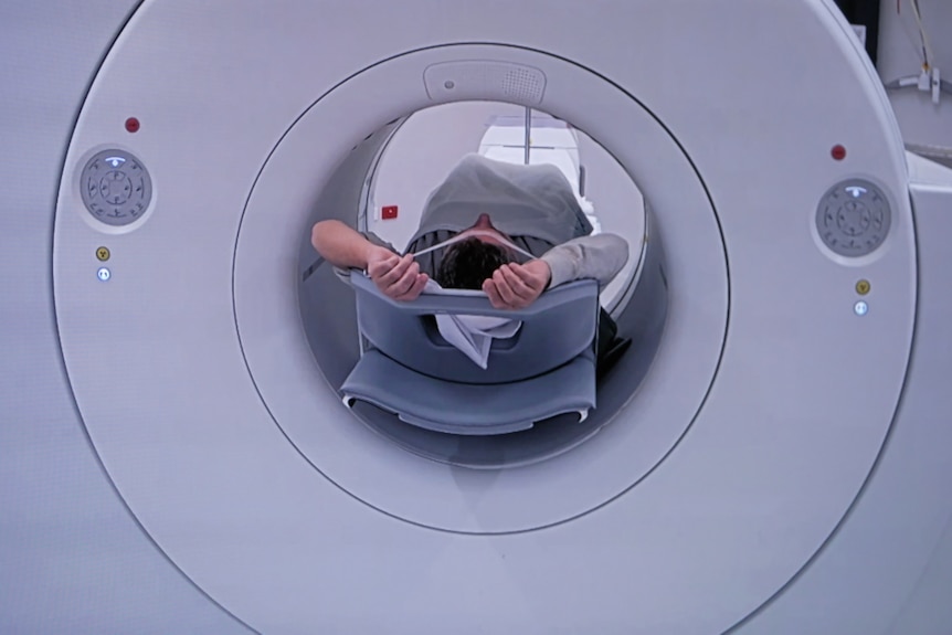 A view down a the middle of a large medical scanner, showing a man with his hands over his head lying on a trolley.