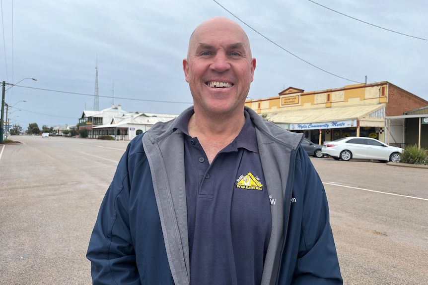 A man in a grey collared t-shirt stands in a street smiling at the camera