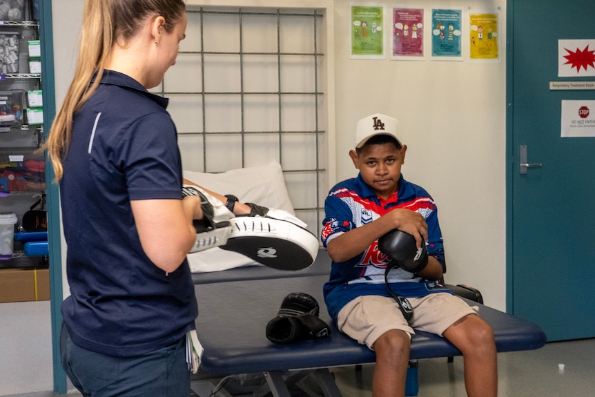 Sam sits and holds a boxing glove while a physio holds some sparring pads