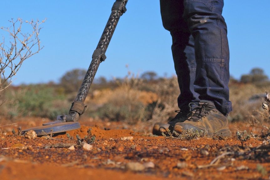 A person waves a metal detector across the red dirt