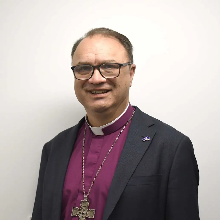 A man wearing a black jacket and purple shirt with an ecclesiastical collar
