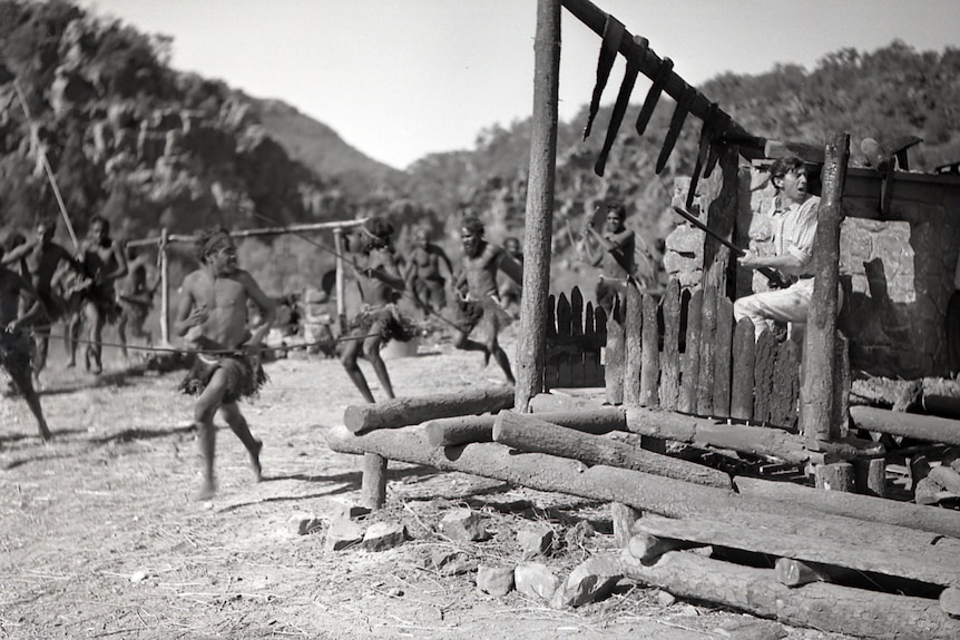 Aboriginal actors running beside a burnt-out ruin of a home, where a male actor stands inside with a rifle.