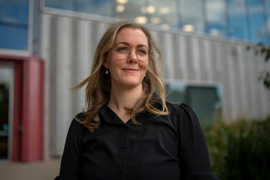 A woman with thin round glasses and blonde hair smiles outside a metallic building with blue sky reflected in windows.