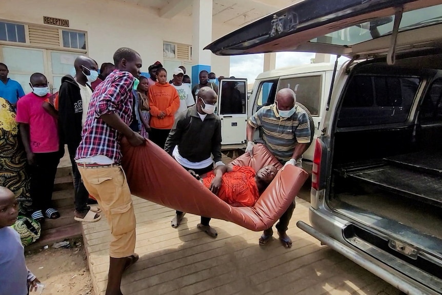 A man with his eyes closed is carried on a mattress into an ambulance with people watching on, wearing masks.