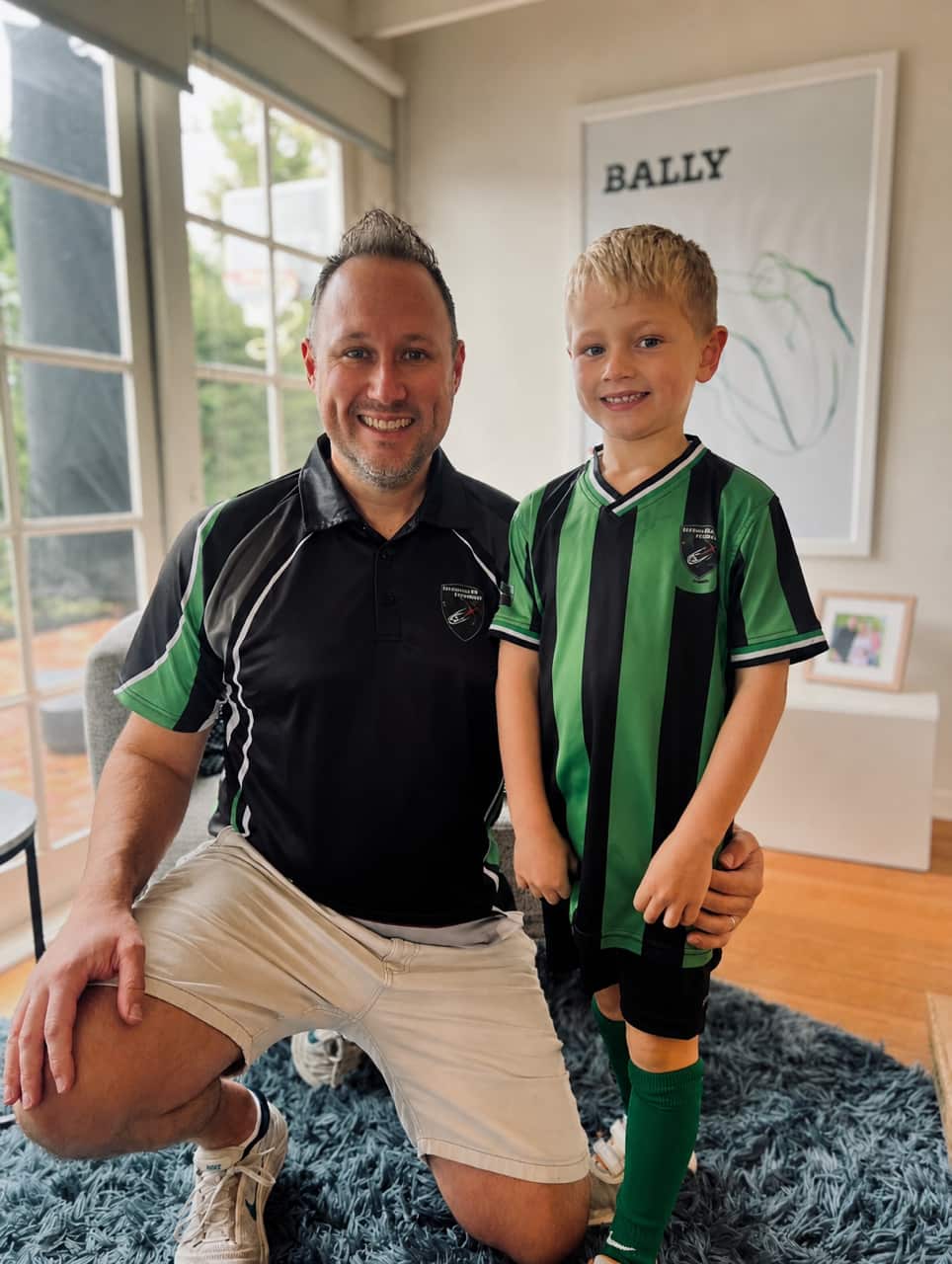 A man is crouched beside a young boy in a room. The man is wearing a black polo shirt with green markings on the sleeves and the boy is wearing a green-and-black striped short-sleeved football shirt