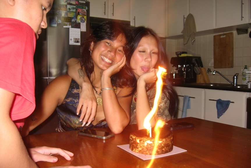 One sister smiling, second sister with tongue out and hoding first sister, behind birthday cake at wooden dinner table